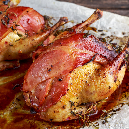 Two roasted partridges wrapped in bacon on a rustic wooden surface.