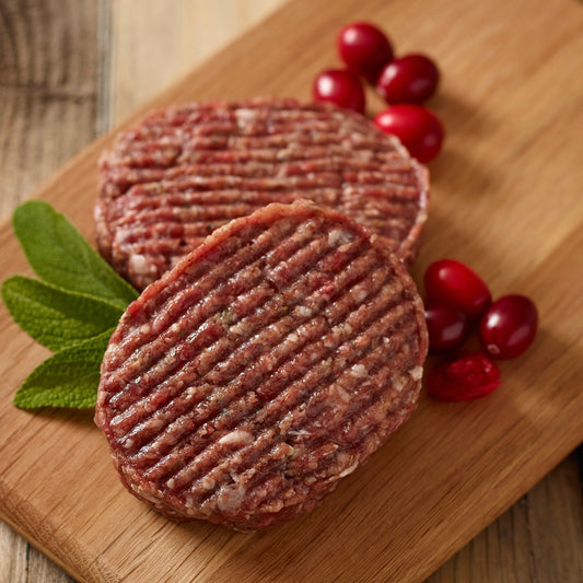 Two raw venison and cranberry hamburger patties on a wooden cutting board with cranberries and mint leaves.