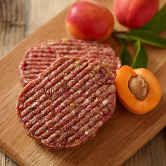 Two raw wild boar and apricot hamburger patties on a wooden cutting board with apricots and leaves.