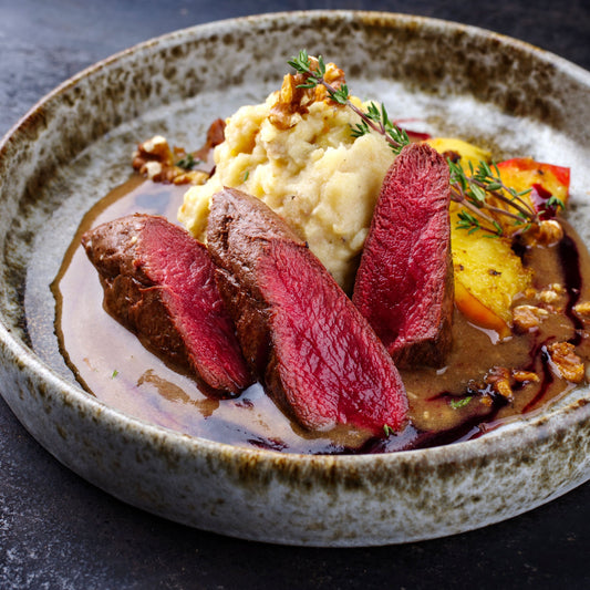 Plated venison haunch game meat dish with sliced red meat, mashed potatoes, and vegetables on a rustic plate.