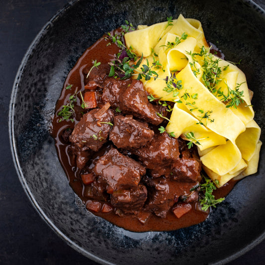 Diced game meat stew with a rich brown sauce served with yellow pasta on a dark plate.
