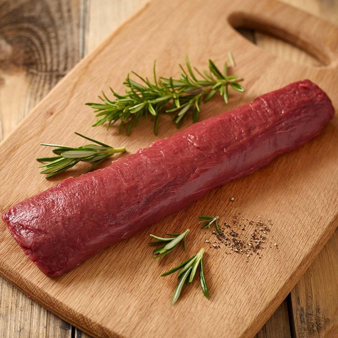 Raw venison striploin on a wooden cutting board with rosemary and pepper.