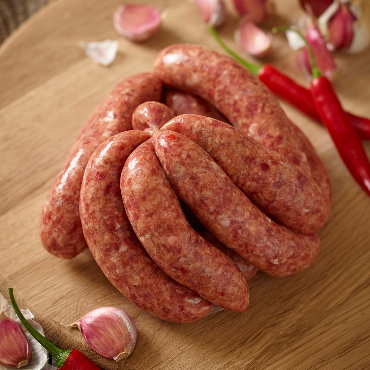 Raw wild boar chilli and garlic sausage link on a wooden board with red peppers and garlic cloves.